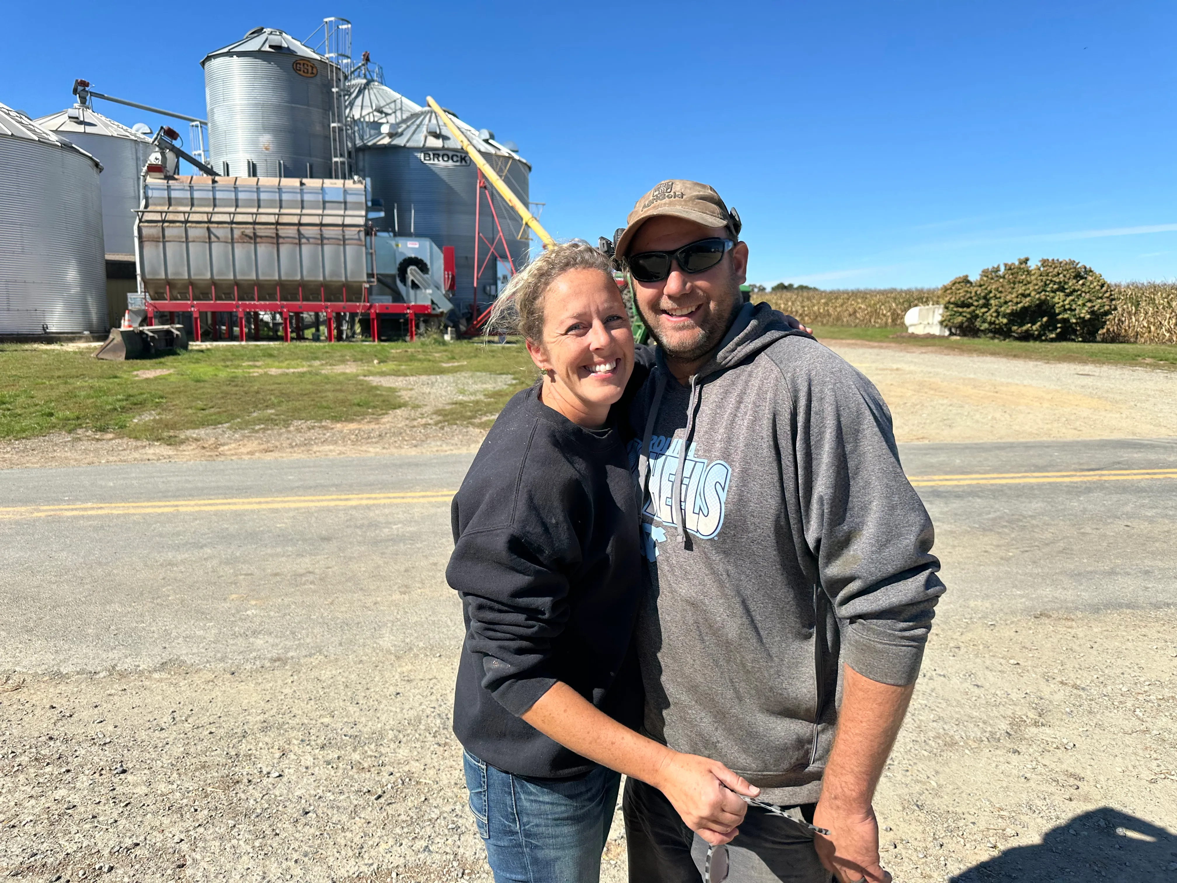 A couple smiles together in front of farm grain silos on a sunny day.